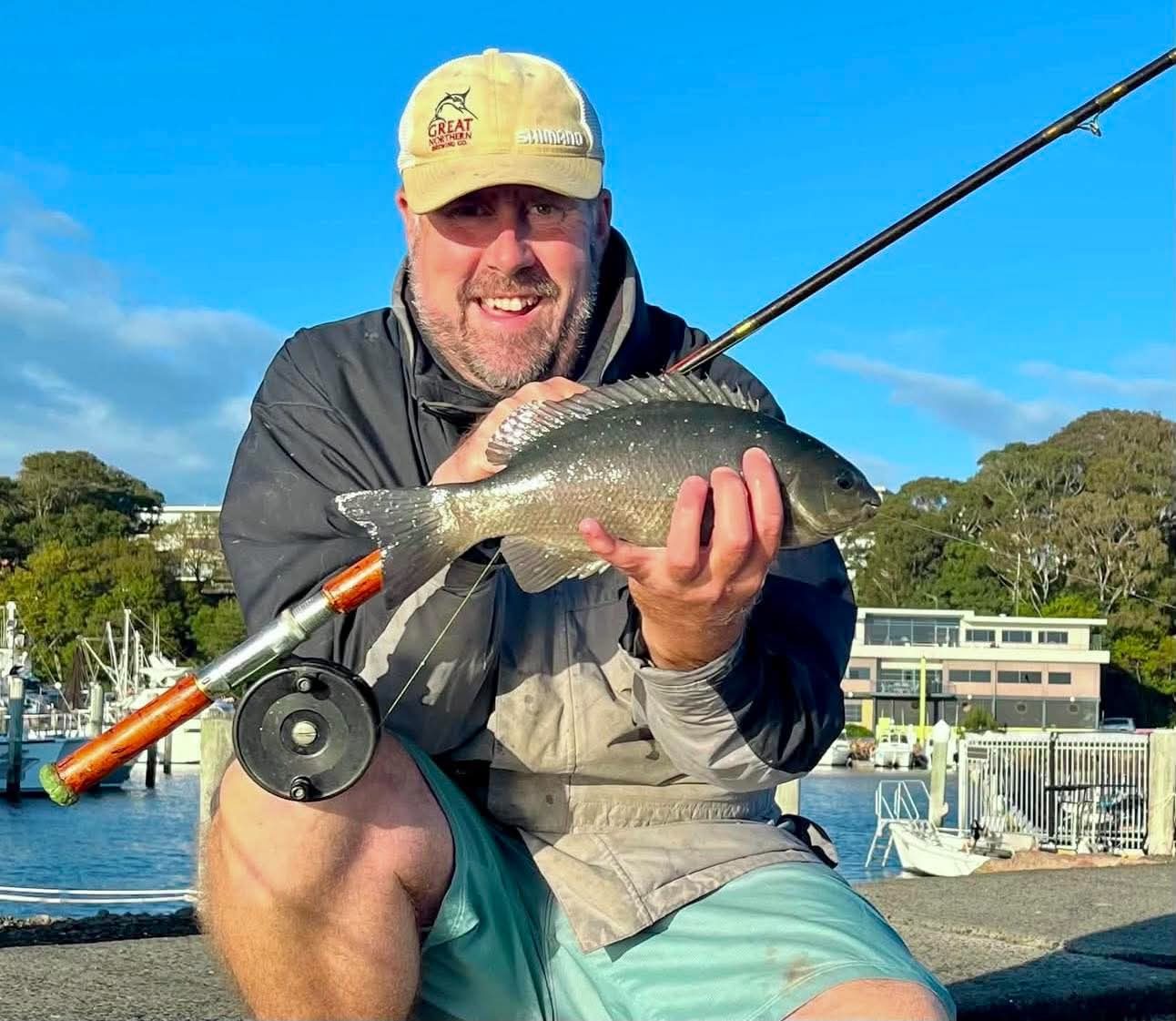 A club member holding a luderick caught from a marina wall, rod and centrepin reel beside him