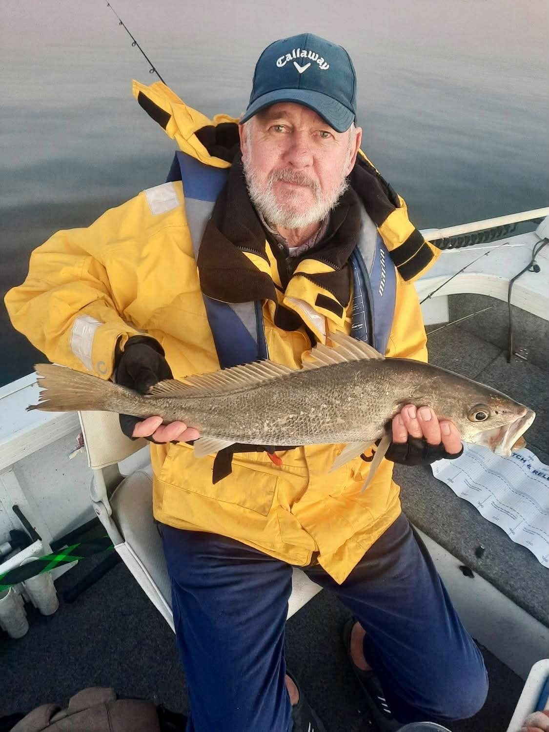 A club member in wet weather gear holding a mulloway on a still, grey morning