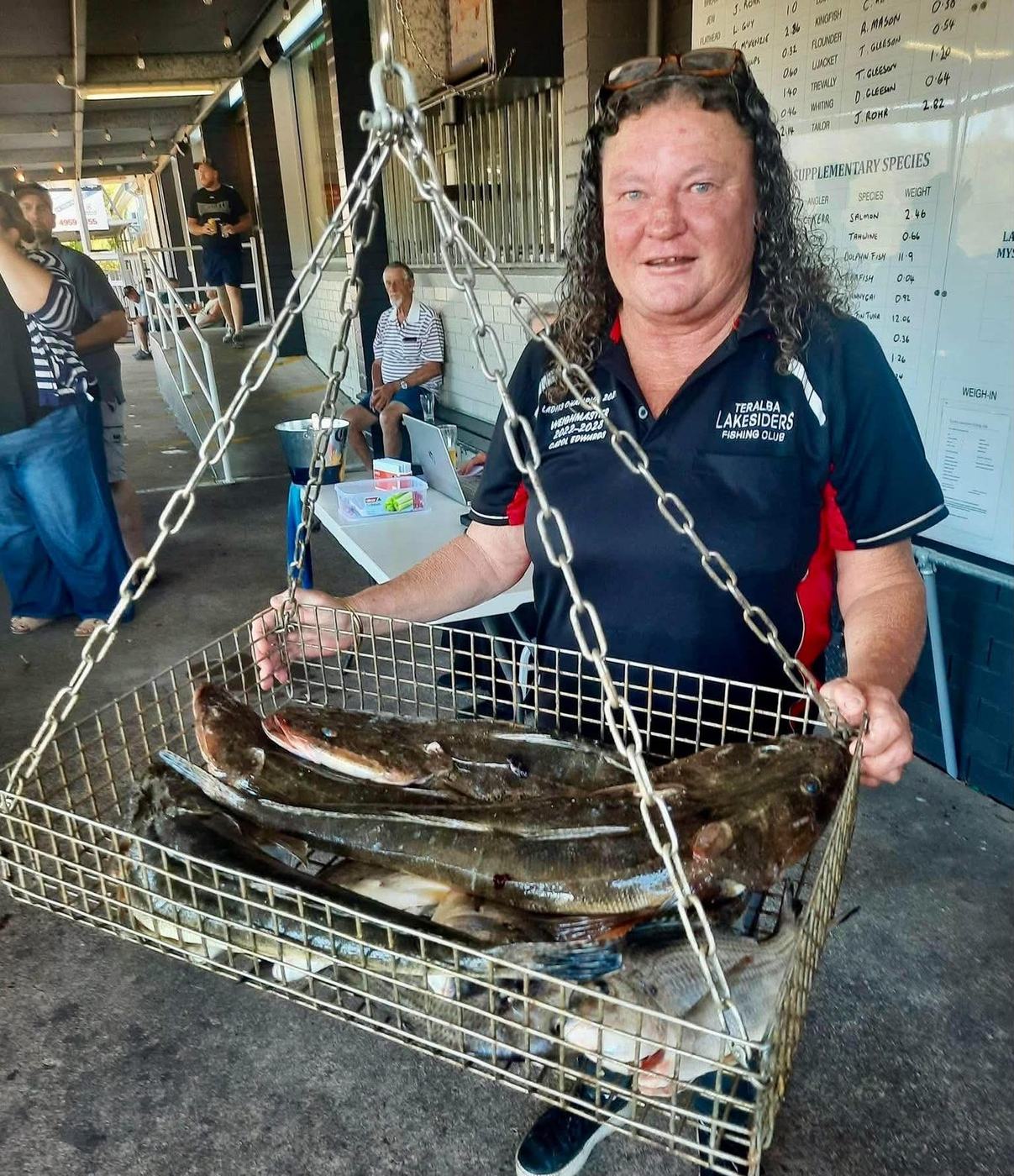 The club weighmaster holding a basket of flathead under the results boards