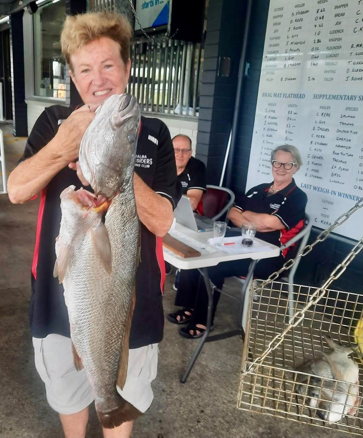 A woman angler holding a large mulloway in front of the club results boards